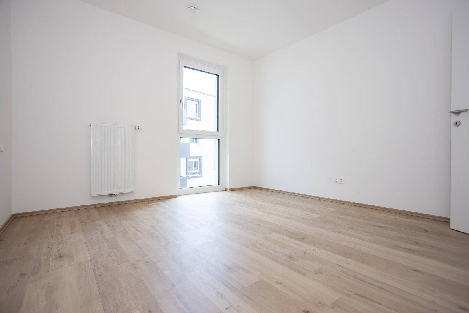 Helles Schlafzimmer mit großem Fenster und hellem Holzboden in einem Neubau.
