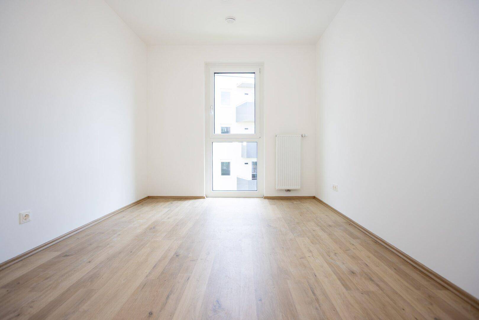 Ruhiges Schlafzimmer mit hellem Holzboden und einem hohen Fenster mit Blick nach draußen.