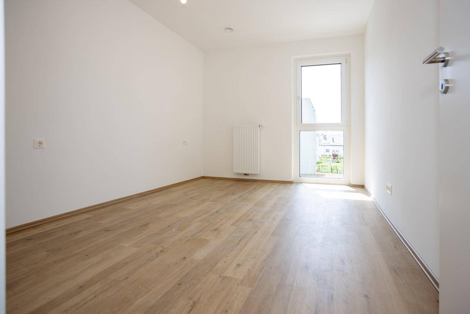 Helles Schlafzimmer mit Laminatboden, weiß gestrichenen Wänden und Zugang zu einem kleinen Balkon.