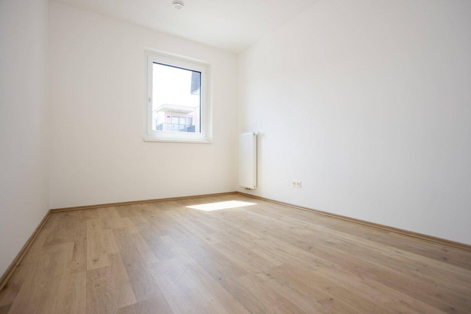 Helles Zimmer mit Laminatboden, weißen Wänden und einem Fenster, das viel Tageslicht hereinlässt.