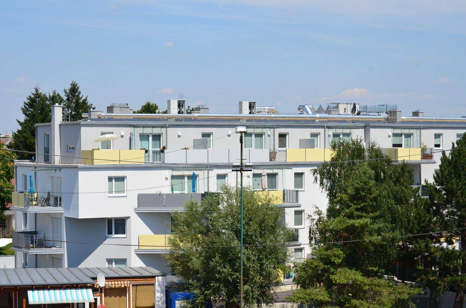 Mehrstöckiges Wohngebäude mit Balkonen und Flachdach, umgeben von Bäumen unter blauem Himmel.