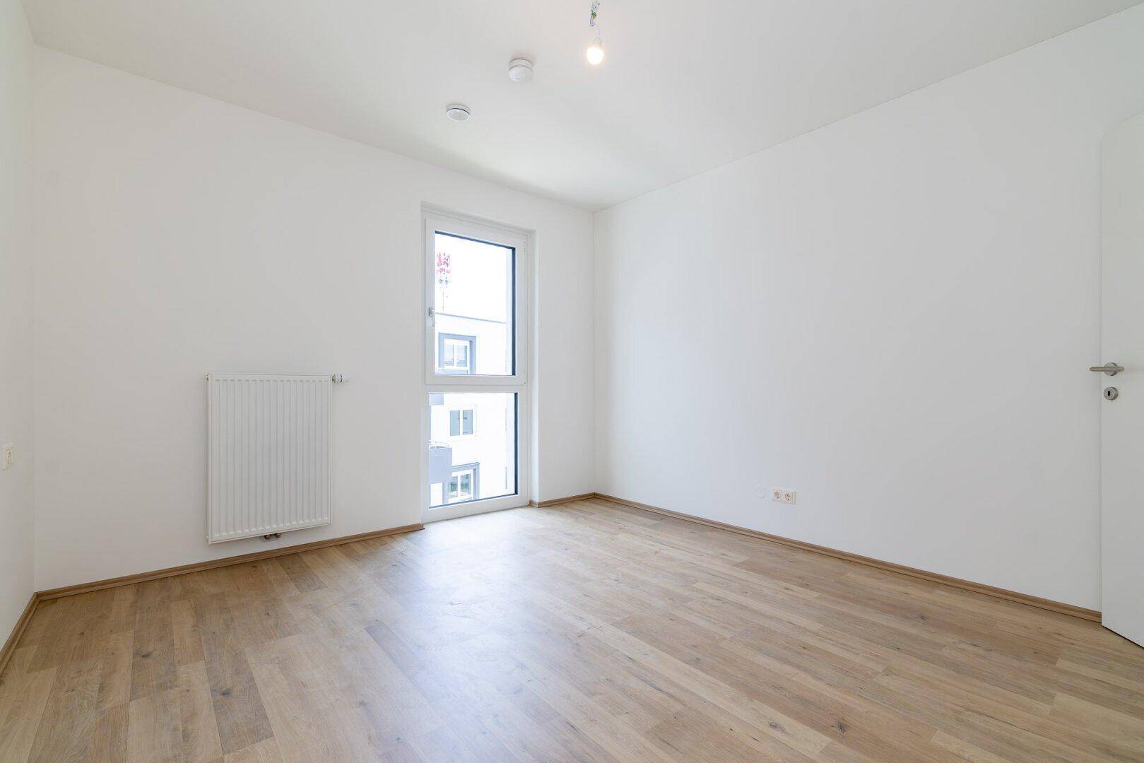 Helles Schlafzimmer mit Laminatboden, weißen Wänden und einem Fenster, das viel Tageslicht bietet.