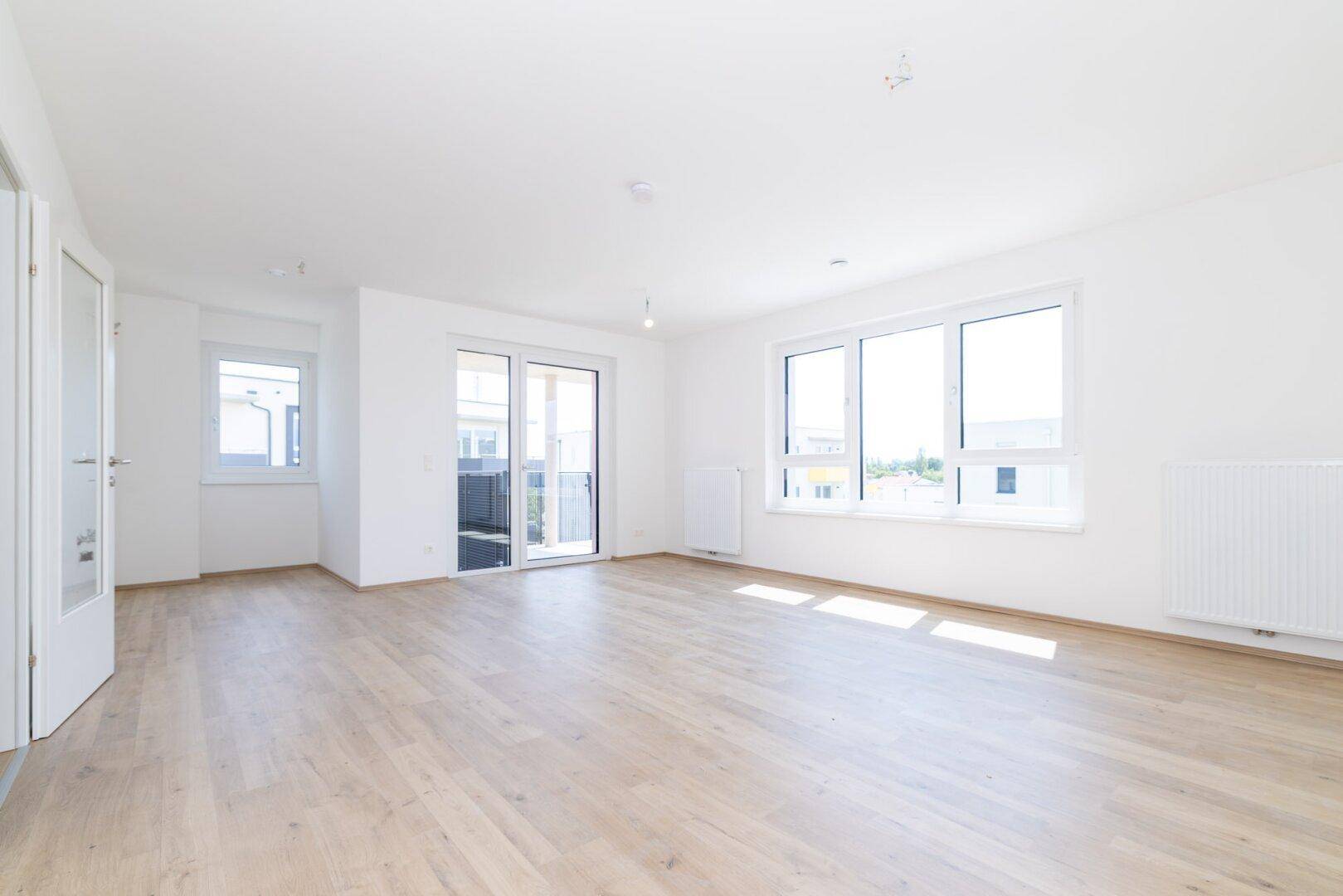 Lichtdurchfluteter Wohnbereich mit hellem Laminatboden, großen Fenstern und Zugang zum Balkon.