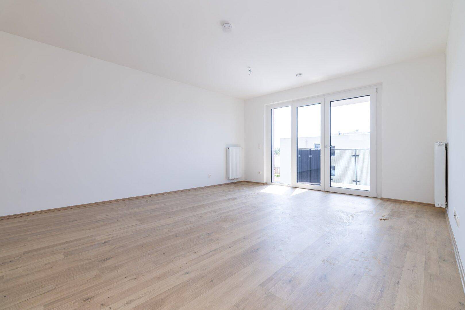 Helles, leeres Zimmer mit hellem Laminatboden, weißen Wänden und Zugang zu einem französischen Balkon.