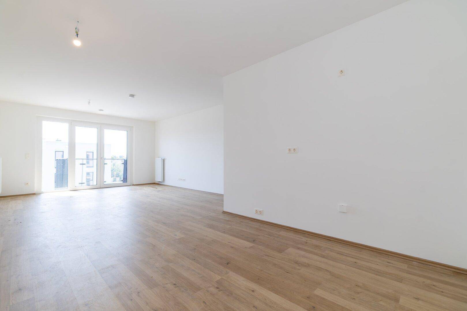 Geräumiges, leeres Zimmer mit Laminatboden, weißen Wänden und viel Tageslicht durch die Balkontür.