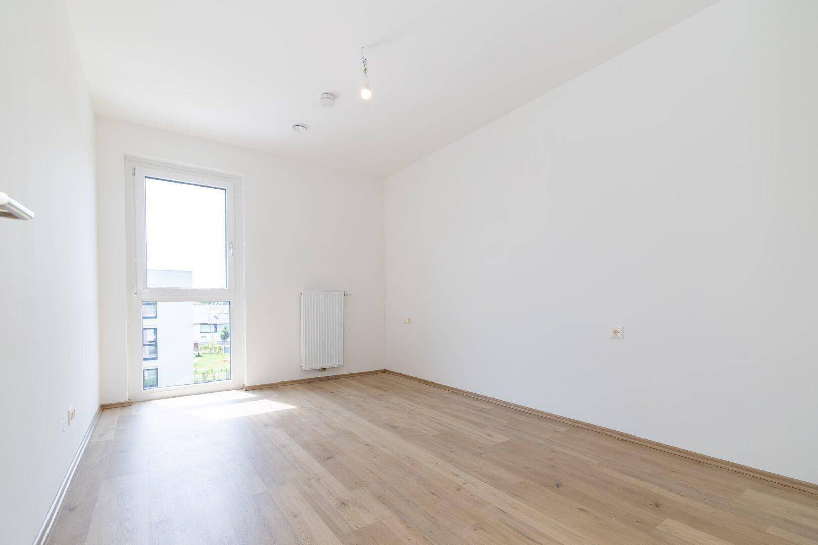 Helles Schlafzimmer mit hellem Laminatboden, weißen Wänden und einem großen Fenster für viel Tageslicht.