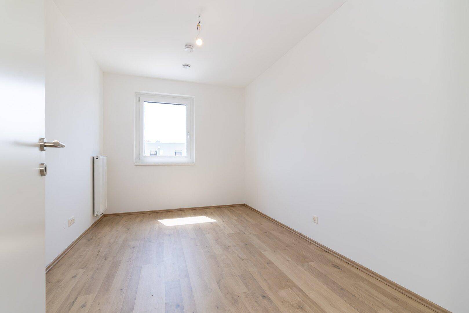 Leeres Schlafzimmer mit hellem Laminatboden, weißen Wänden und einem Fenster, das Sonnenlicht hereinlässt.