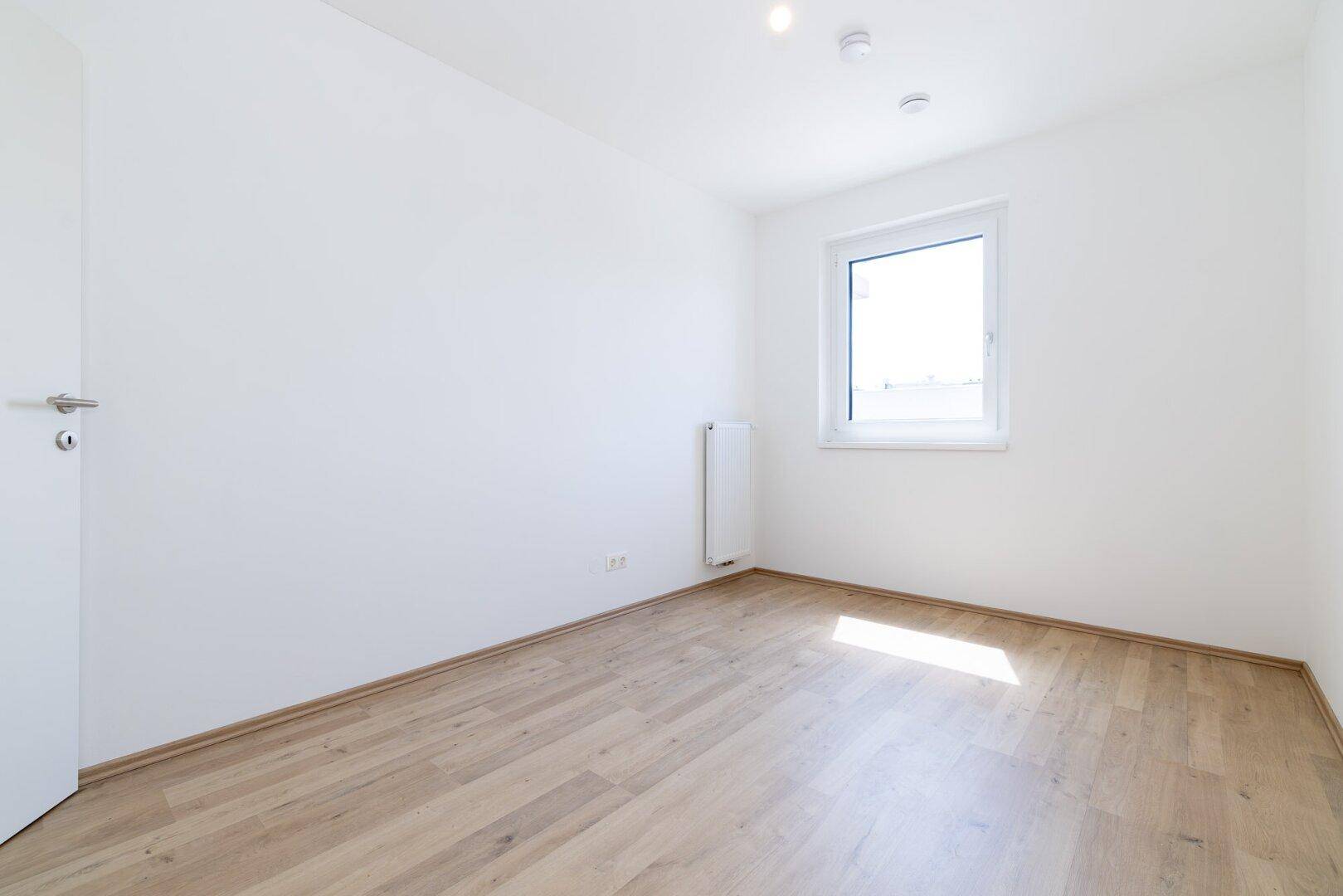 Helles, leeres Zimmer mit hellem Laminatboden, weißen Wänden und einem Fenster, das viel Licht spendet.