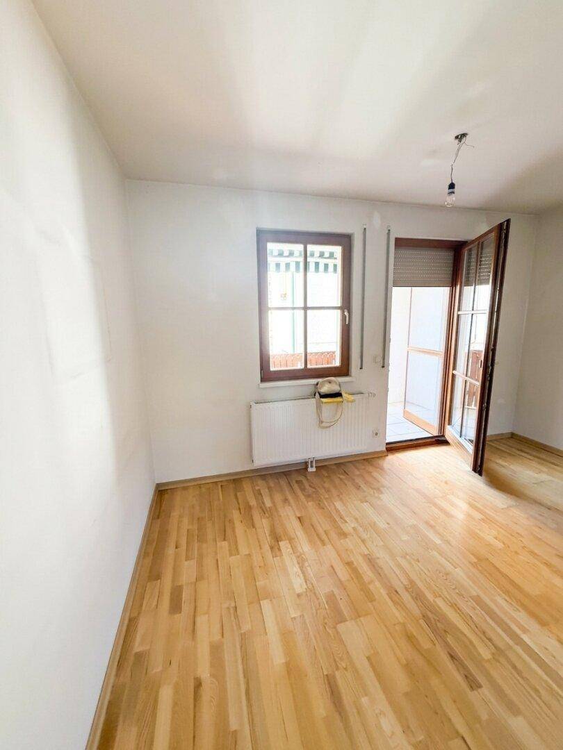 Heller, leerer Raum mit Holzboden, Fenster und Zugang zu einem Außenbereich.