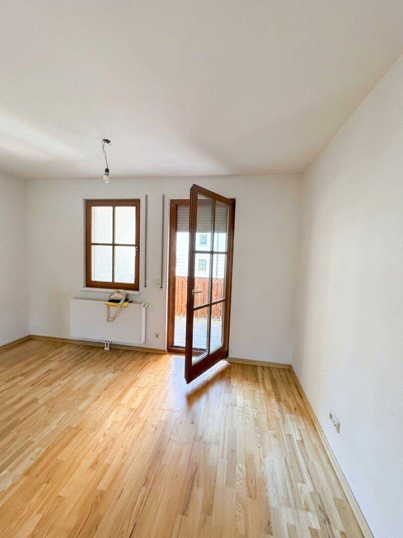 Geräumiger, unmöblierter Raum mit hellem Holzboden und Zugang zum Balkon.