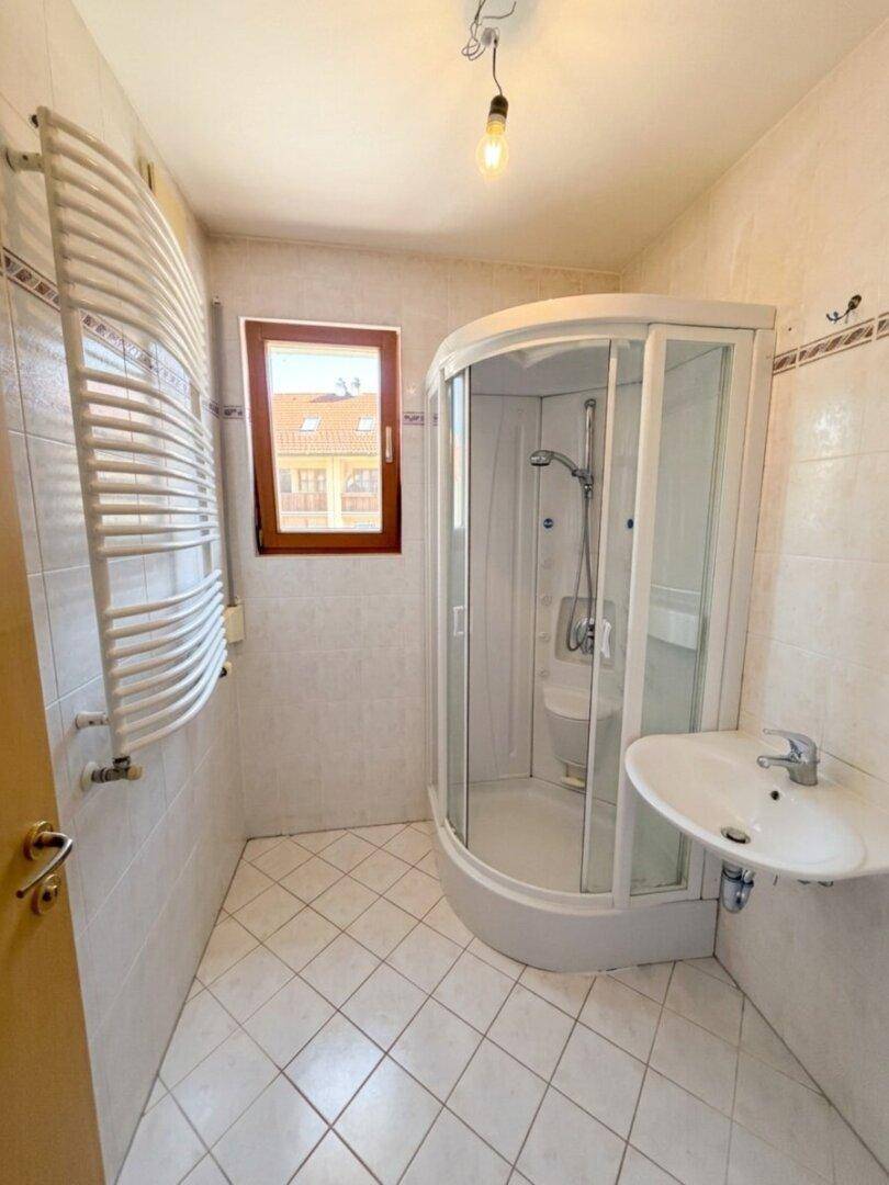 Helles Badezimmer mit Eckduschkabine, Waschbecken, Handtuchheizkörper und Fenster.