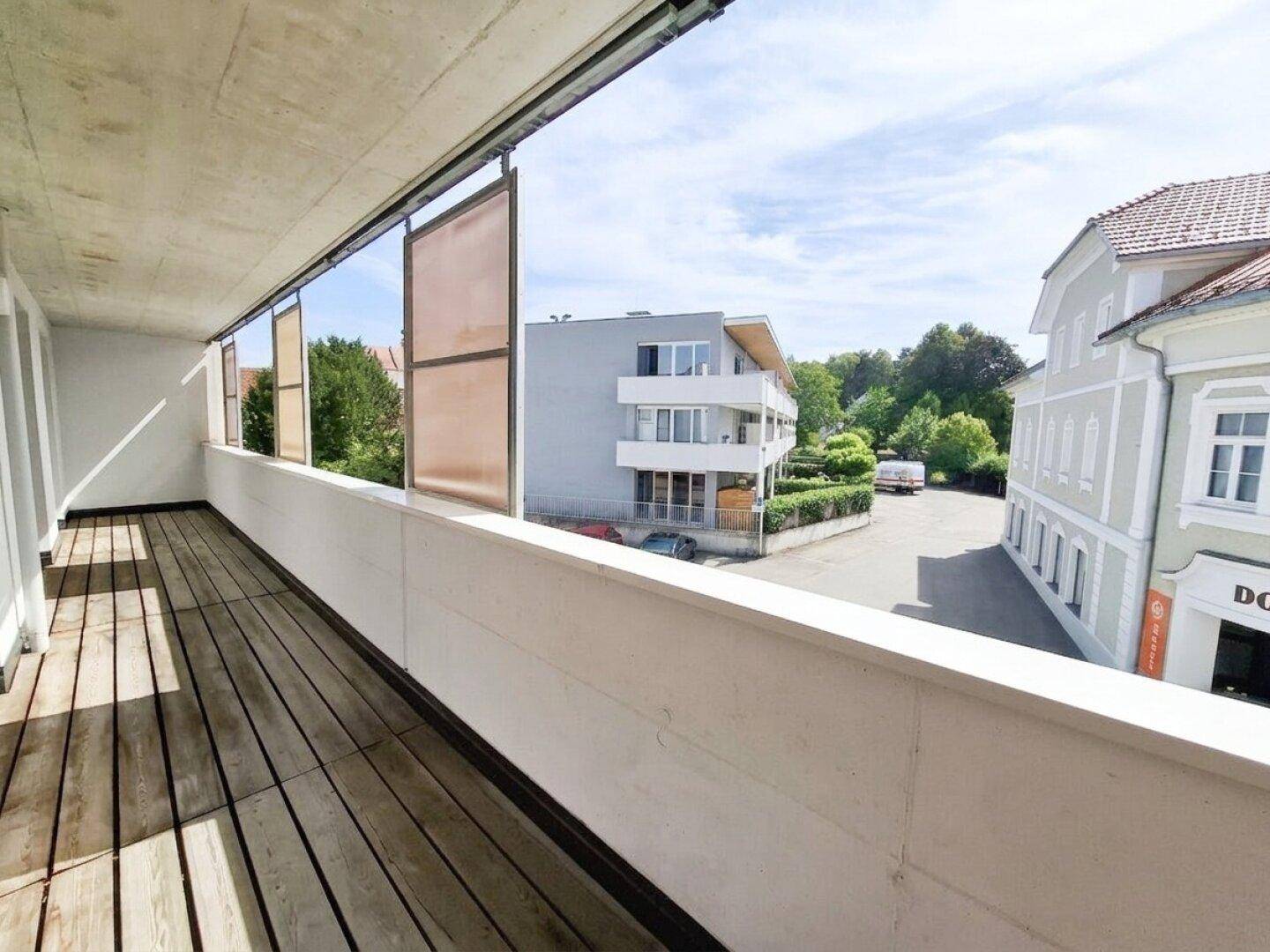 Langer Balkon mit Holzboden und Blick auf die umliegenden Gebäude und grüne Umgebung.