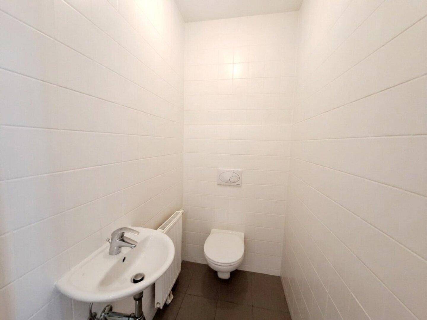 Separates WC mit wandhängender Toilette und kleinem Waschbecken, hell gefliest.