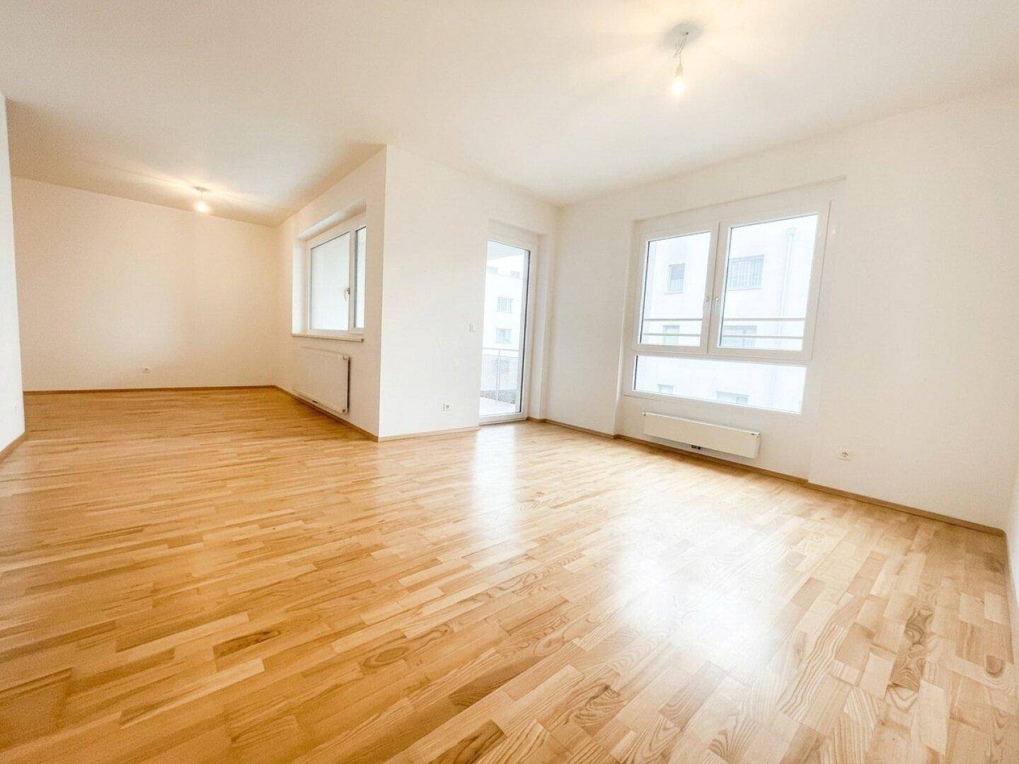 Lichtdurchfluteter Wohnbereich mit Parkettboden, großen Fenstern und Zugang zu einem Balkon.