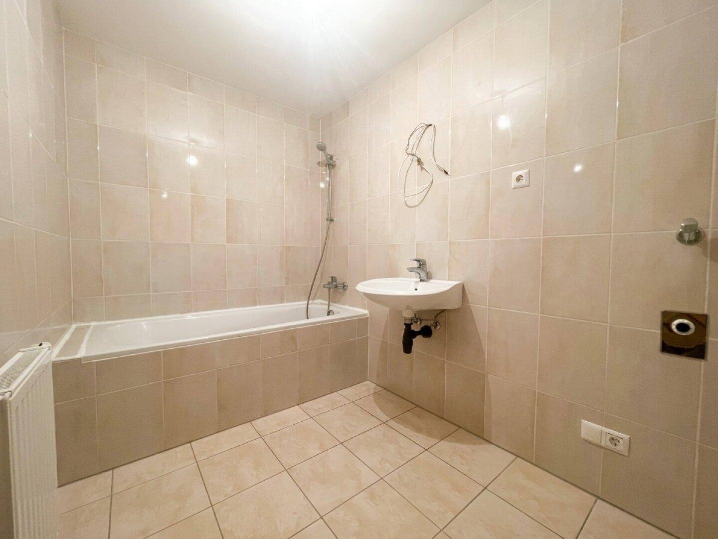 Gefliestes Badezimmer mit Badewanne, Duschfunktion und Waschbecken, in neutralen Beigetönen gehalten.