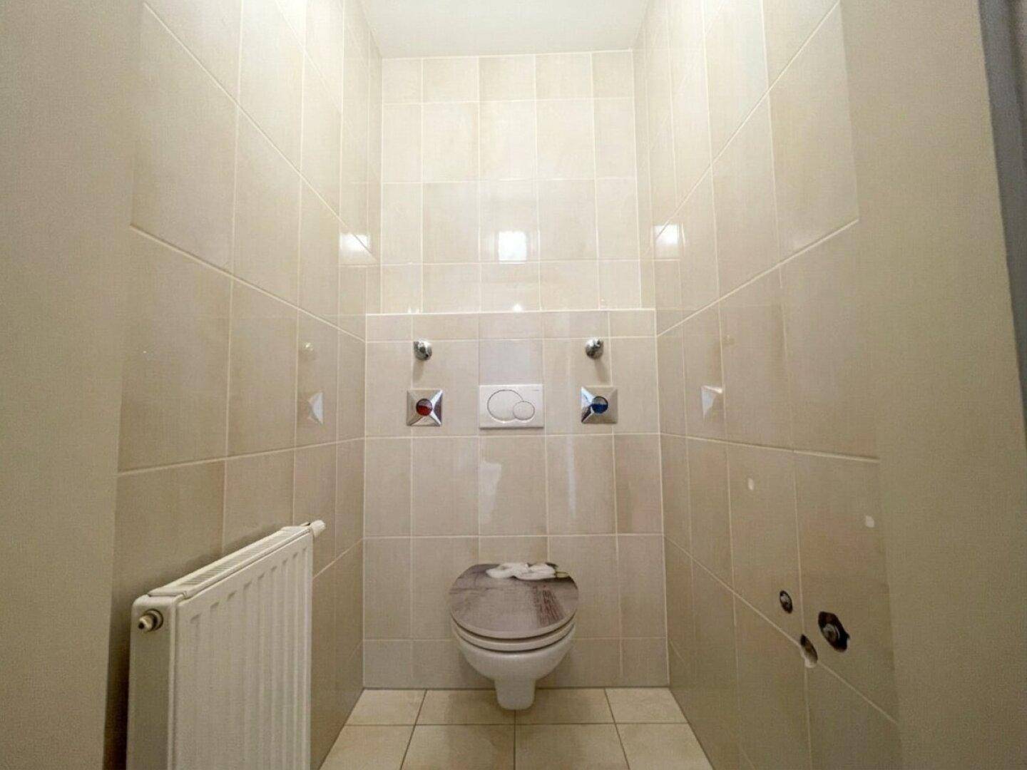 Separates WC, komplett gefliest mit wandhängender Toilette und Heizkörper, in hellen Beigetönen.