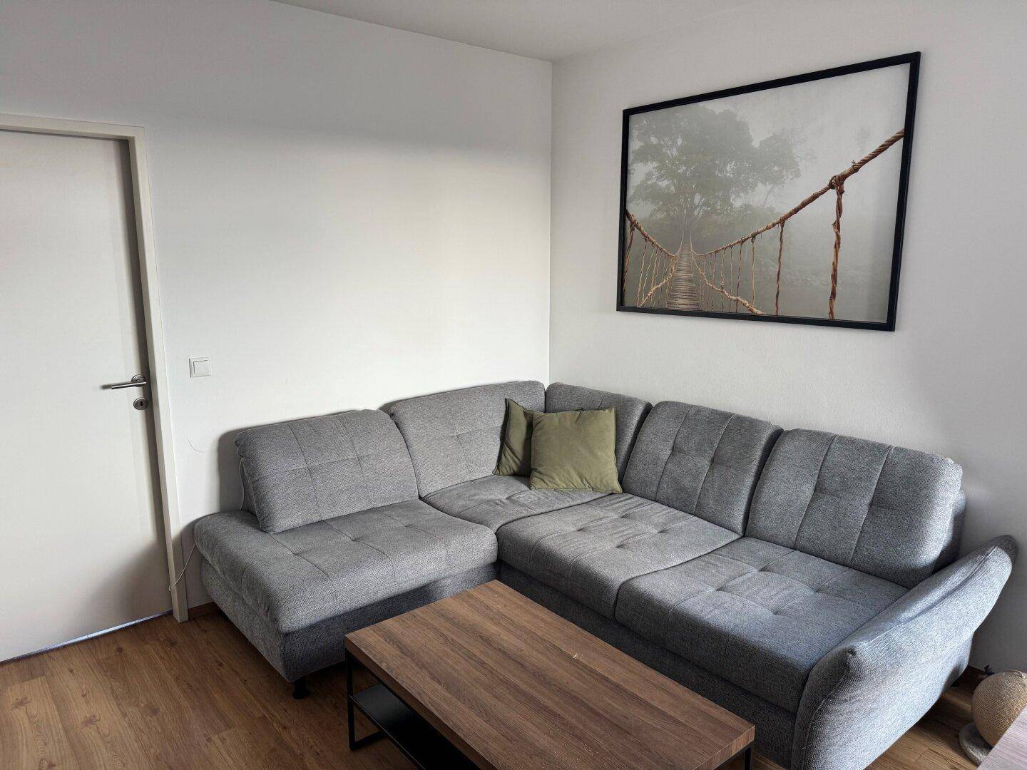 Gemütliches Wohnzimmer mit grauem Ecksofa, Holztisch und einem Bild an der weißen Wand.