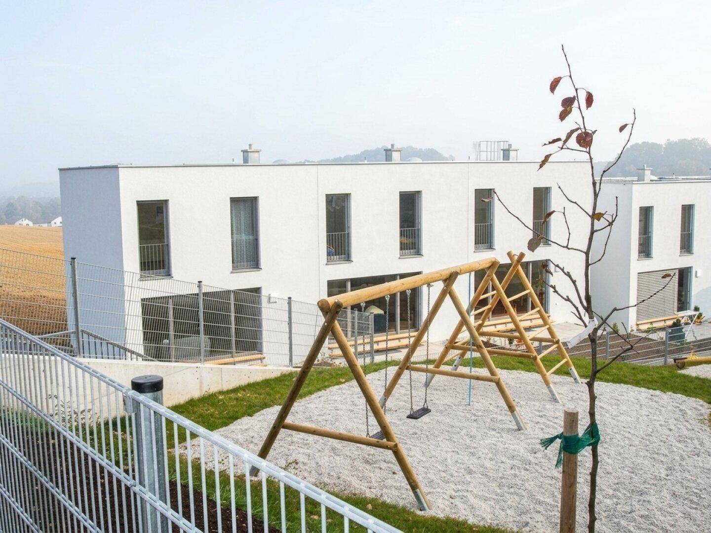 Spielplatz mit Schaukeln und Sandfläche vor einem modernen Wohngebäude, umgeben von Natur.