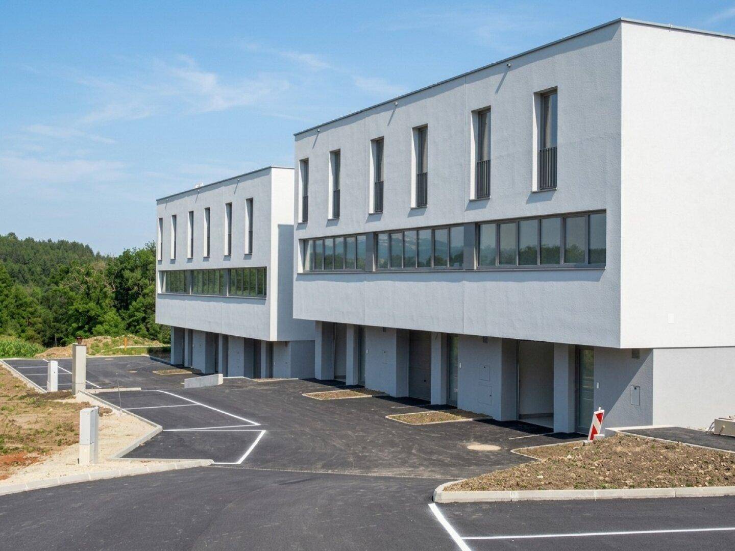 Moderne weiße Gebäude mit großen Fenstern und einem neu angelegten Parkplatz unter blauem Himmel.