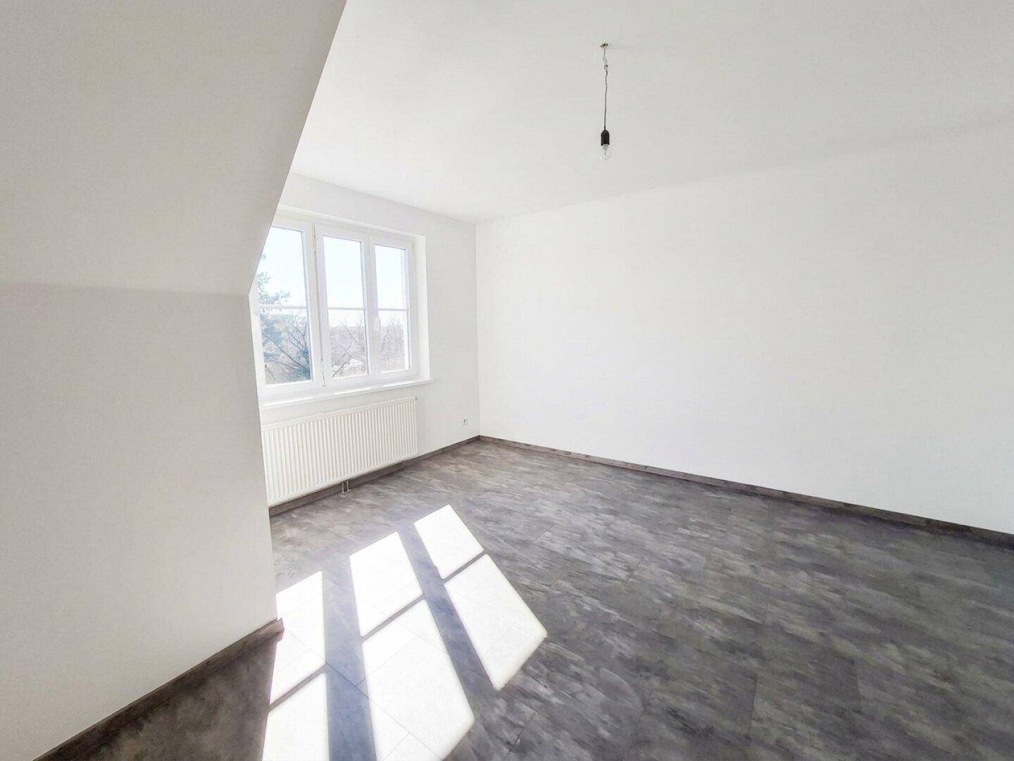 Heller, leerer Raum mit weißen Wänden, großem Fenster und dunklem Fliesenboden.