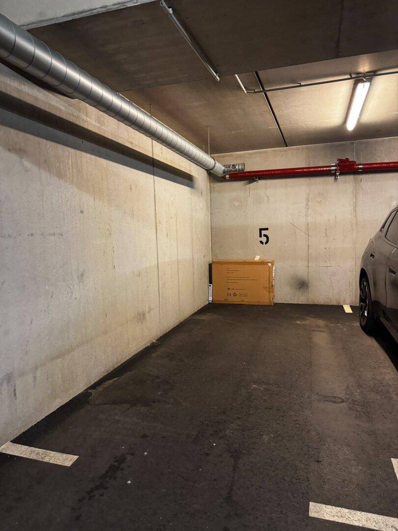 Leerer Tiefgaragenstellplatz mit Betonwänden, Lüftungsrohren und einer Kiste an der Wand platziert.