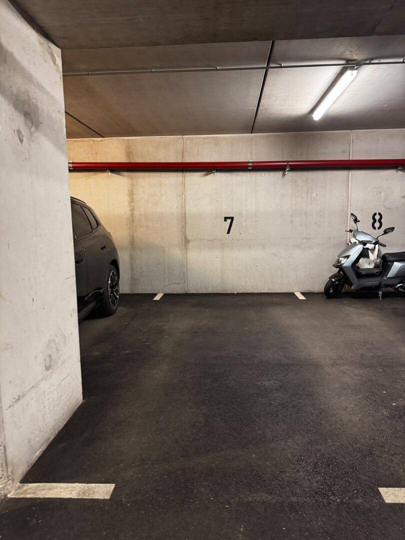 Tiefgaragenstellplatz mit Betonpfeiler, roten Leitungen und einem Roller am Rand des Stellplatzes.