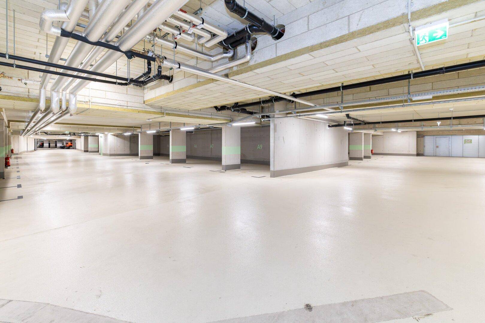 Moderne Tiefgarage mit vielen Stellplätzen, sichtbaren Rohren und heller Beleuchtung.