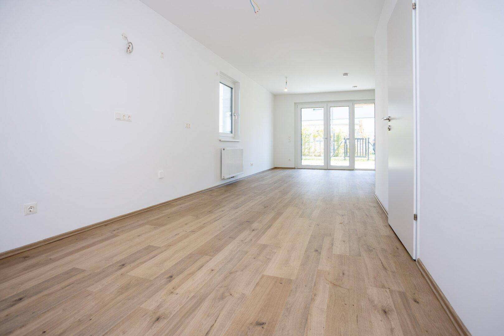 Heller, leerer Wohnraum mit modernem Laminatboden, weißen Wänden und Zugang zur Terrasse.