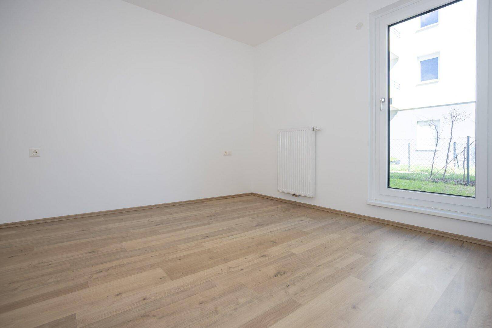 Helles Schlafzimmer mit Laminatboden, weiß gestrichenen Wänden und großem Fenster mit Grünblick.