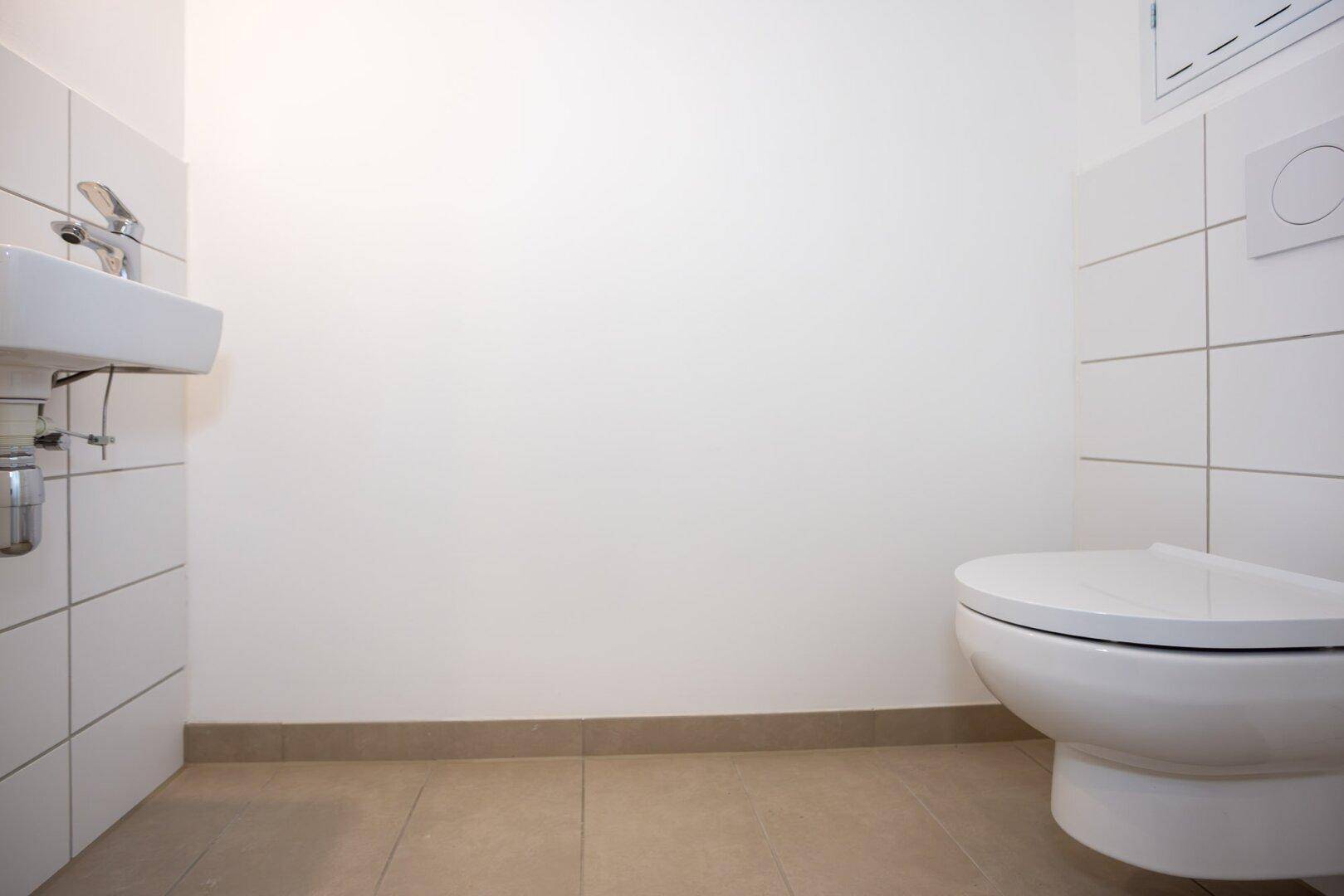 Separates WC mit wandhängender Toilette, kleinem Waschbecken und weißen Fliesen.