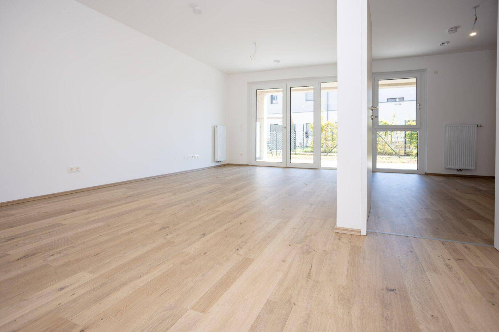 Helles, leeres Wohnzimmer mit hellem Holzboden und großen Fenstern zum Außenbereich.
