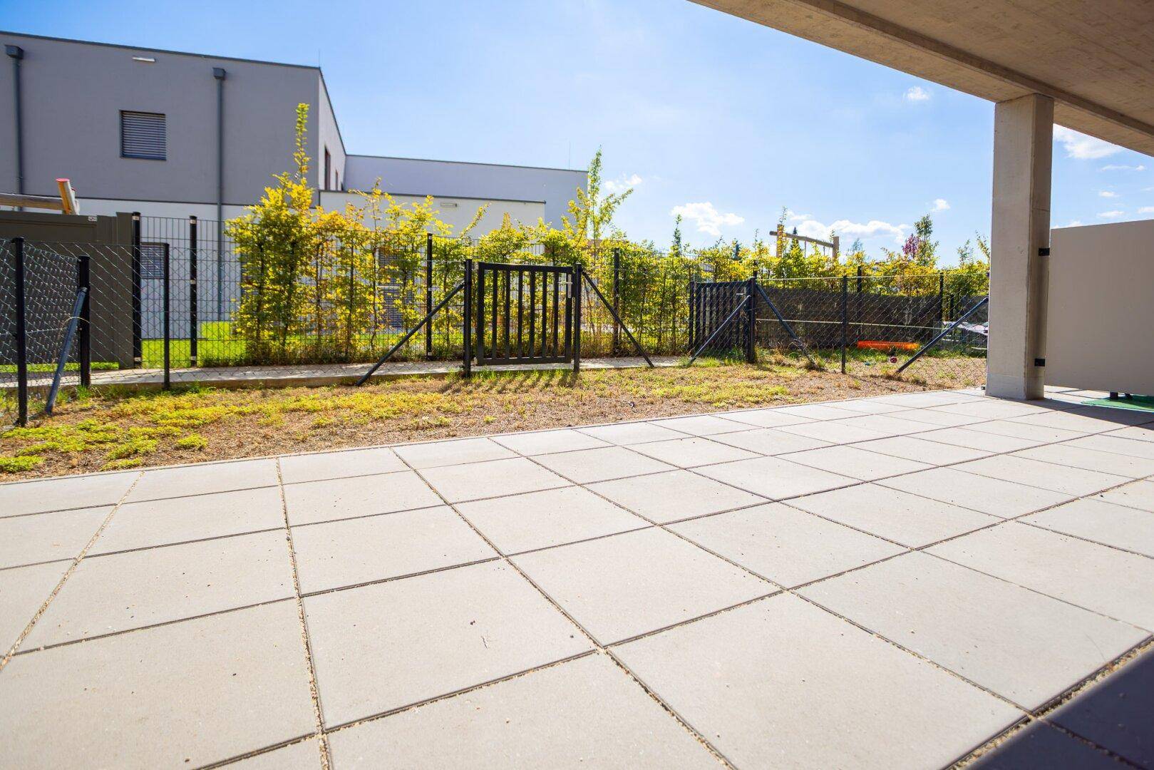 Gepflasterte Terrasse mit Blick auf einen umzäunten Bereich und angrenzende moderne Gebäude.
