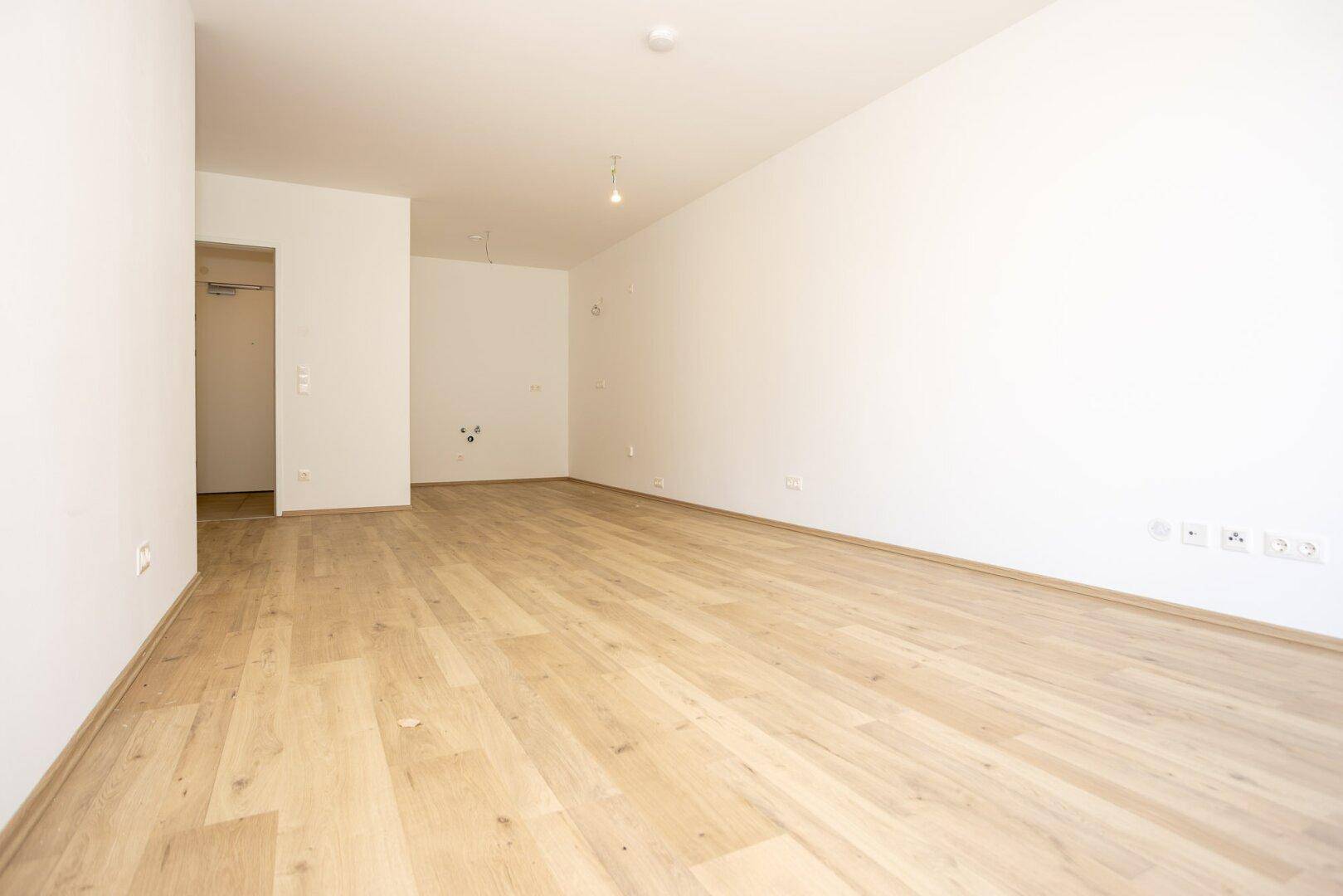 Geräumiger, unmöblierter Raum mit hellem Holzboden und Steckdosen an den weißen Wänden.