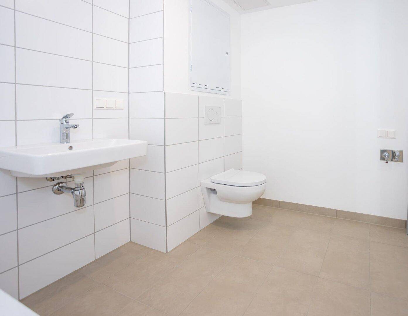 Helles Badezimmer mit weißen Wandfliesen, Waschbecken und wandhängender Toilette.