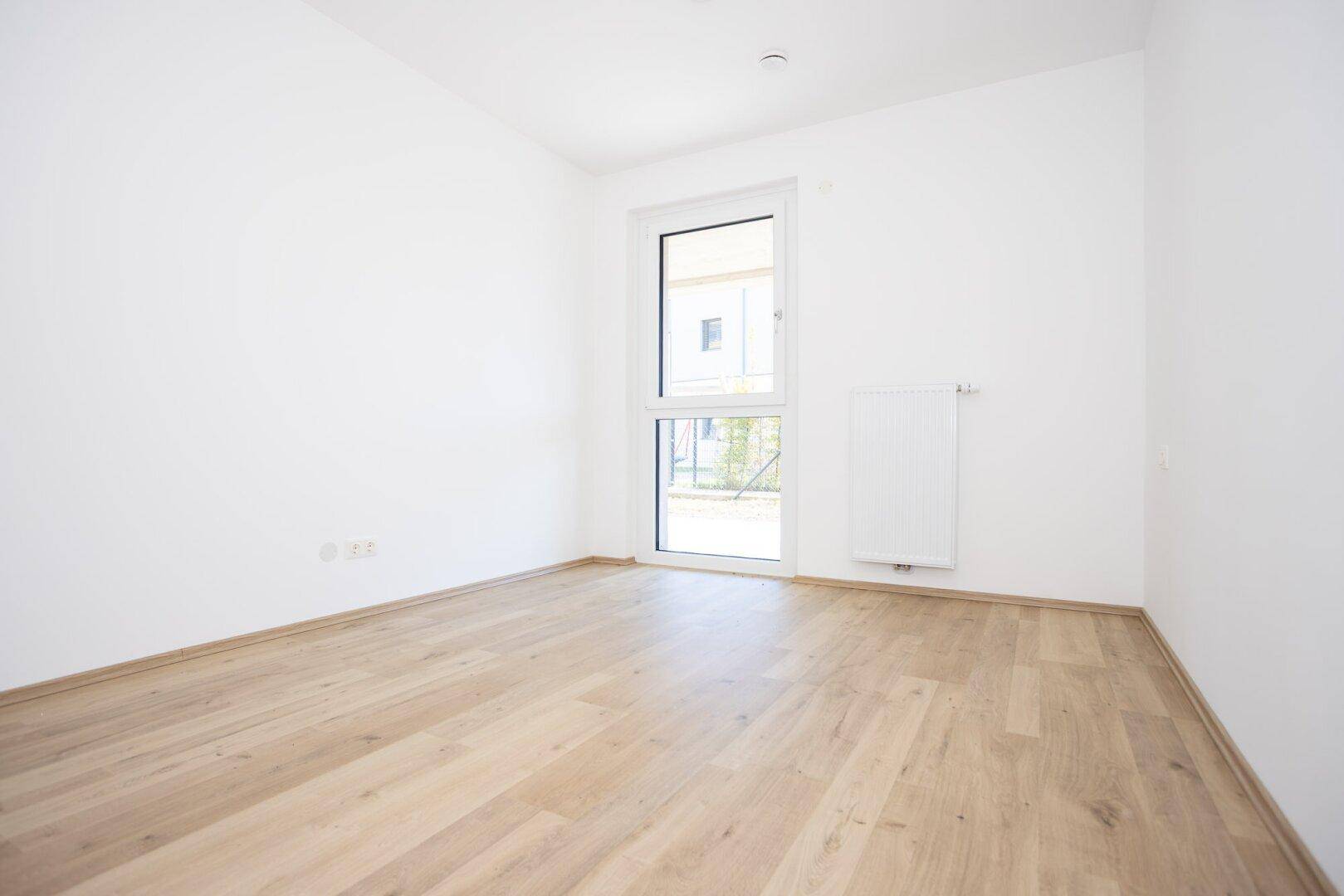 Leeres Schlafzimmer mit hellem Holzboden und einem hohen Fenster, das viel Tageslicht hereinlässt.