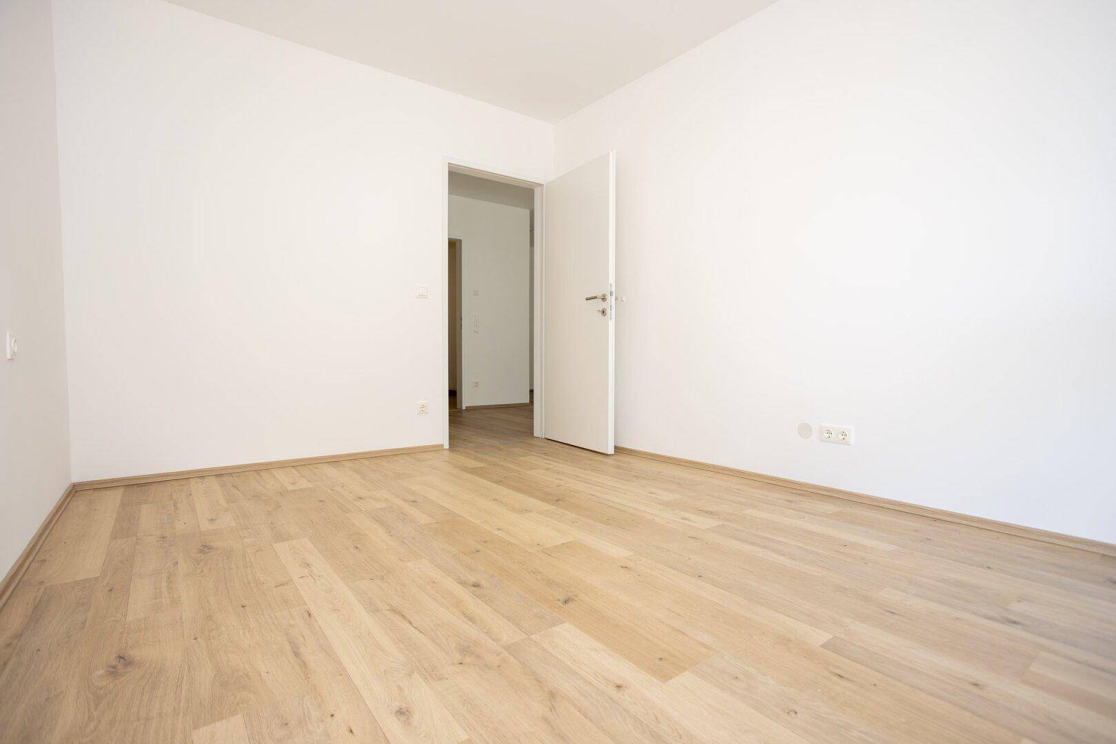 Unmöbliertes Schlafzimmer mit Holzboden und weißer Tür, die zu einem anderen Raum führt.