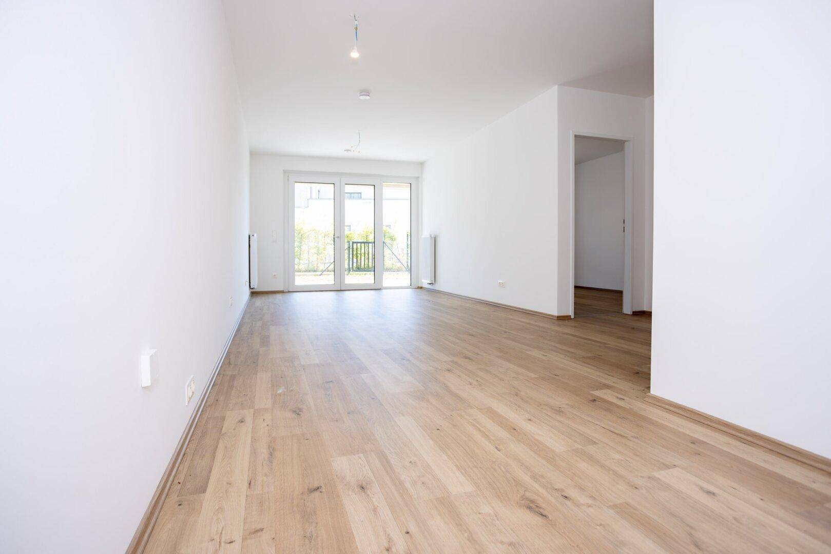 Lichtdurchfluteter Wohnbereich mit hellem Holzboden und großen Fenstertüren, die nach draußen führen.