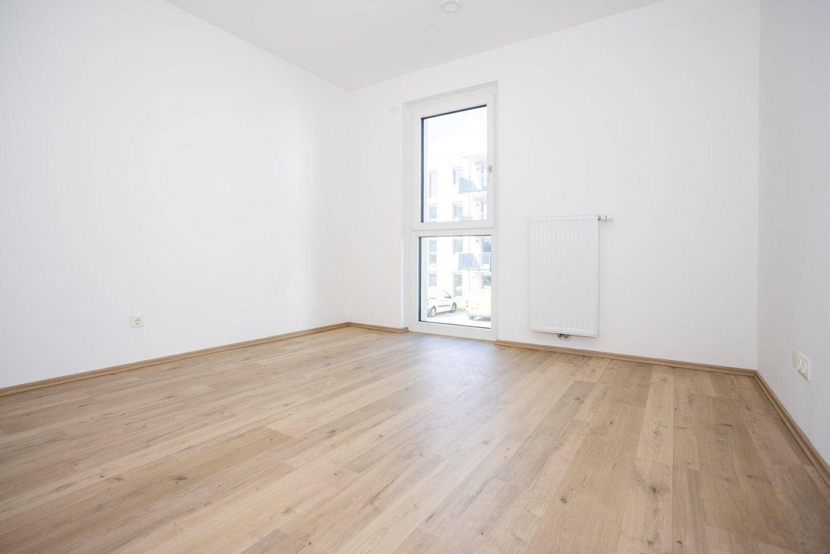 Helles Schlafzimmer mit Laminatboden und großem Fenster, das viel Tageslicht hereinlässt.