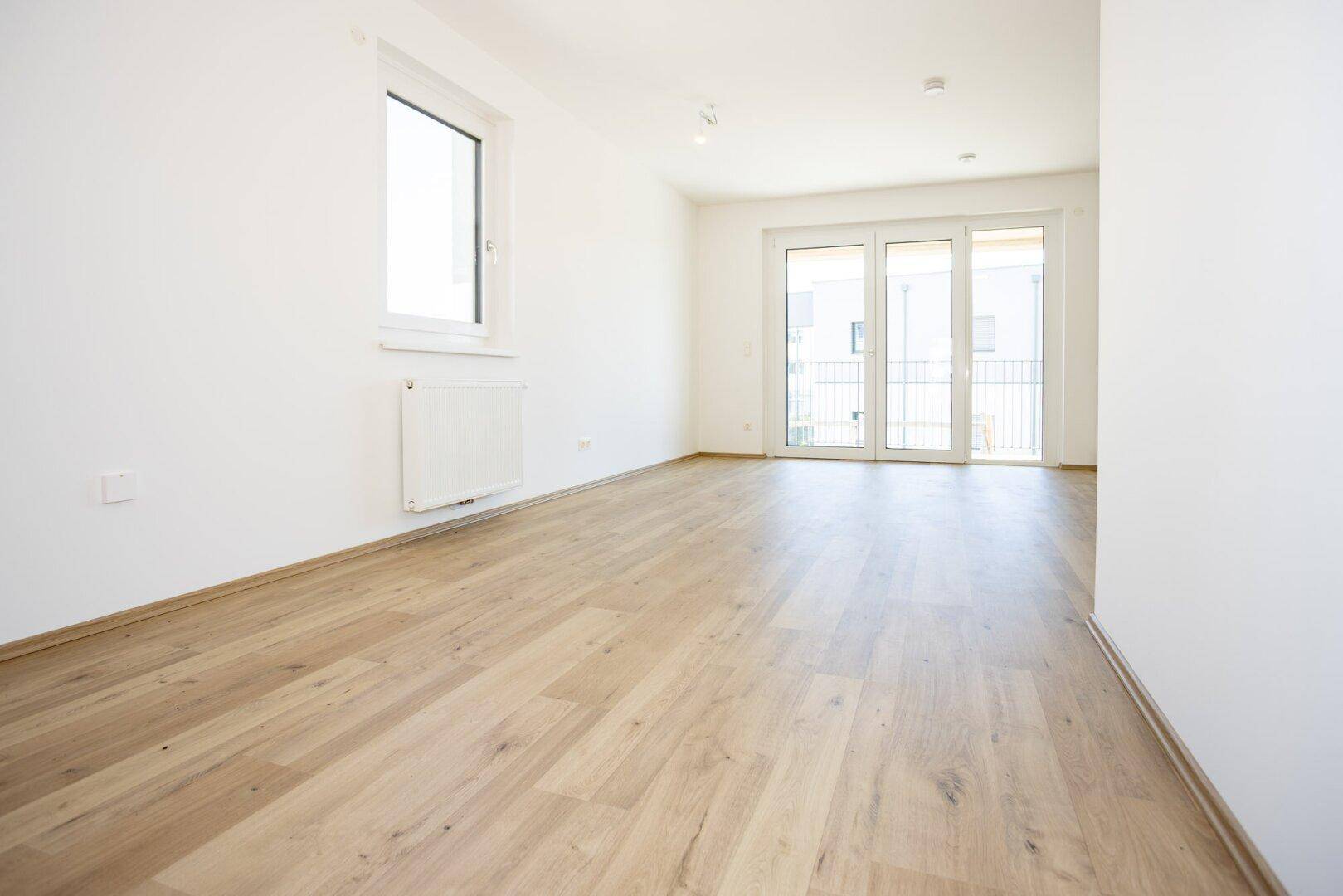 Helles, leeres Zimmer mit hellem Laminatboden, weißen Wänden und Zugang zu einem französischen Balkon.