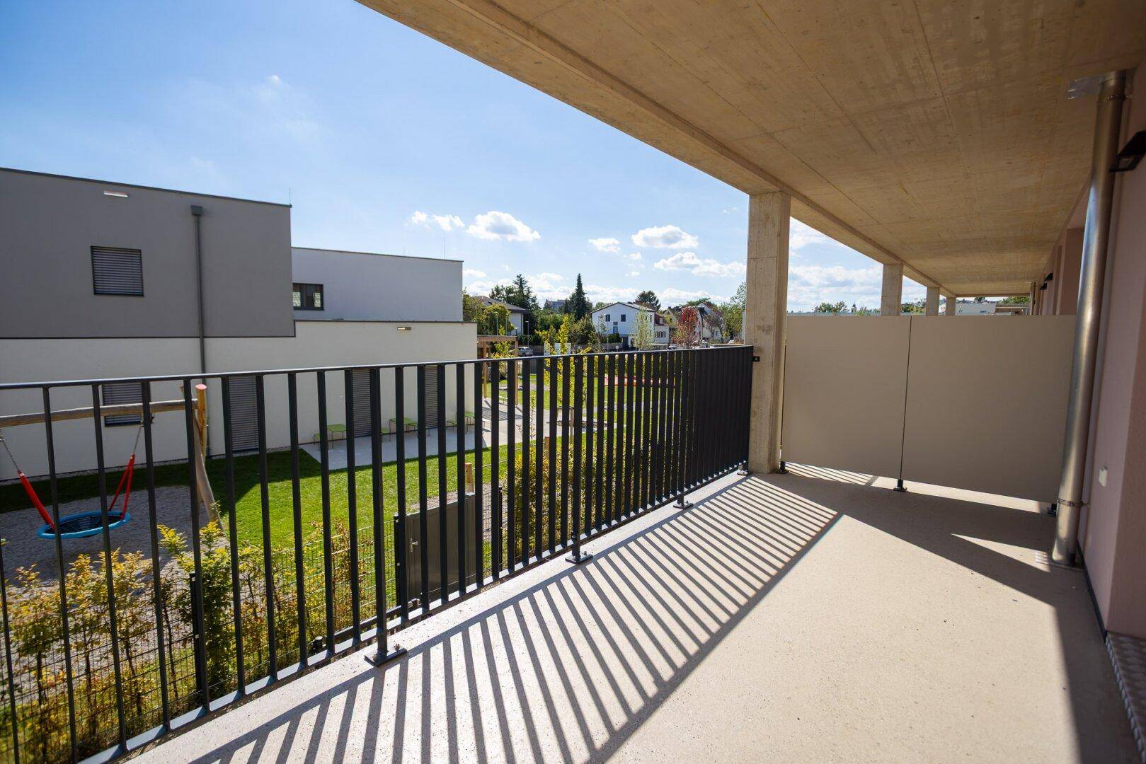 Heller Balkon mit Blick auf den Spielplatz und die umliegenden Wohngebäude an einem sonnigen Tag.