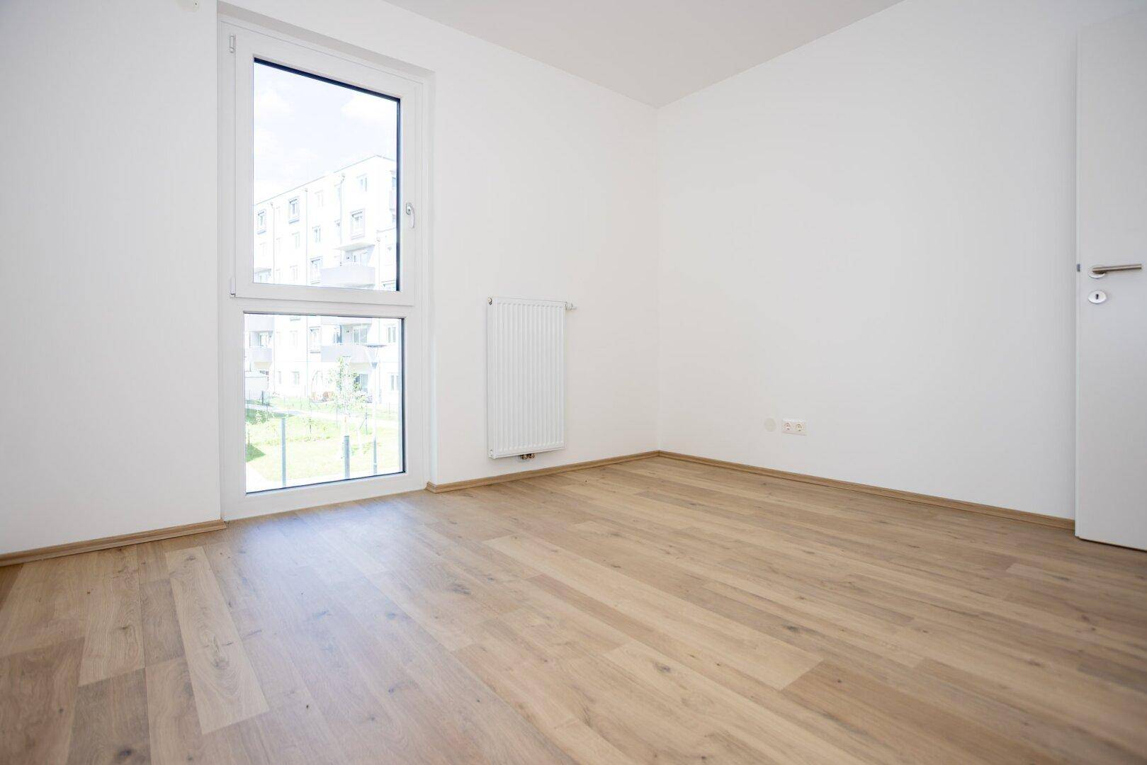 Unmöbliertes Schlafzimmer mit hellem Laminatboden und einem großen Fenster mit Blick ins Grüne.