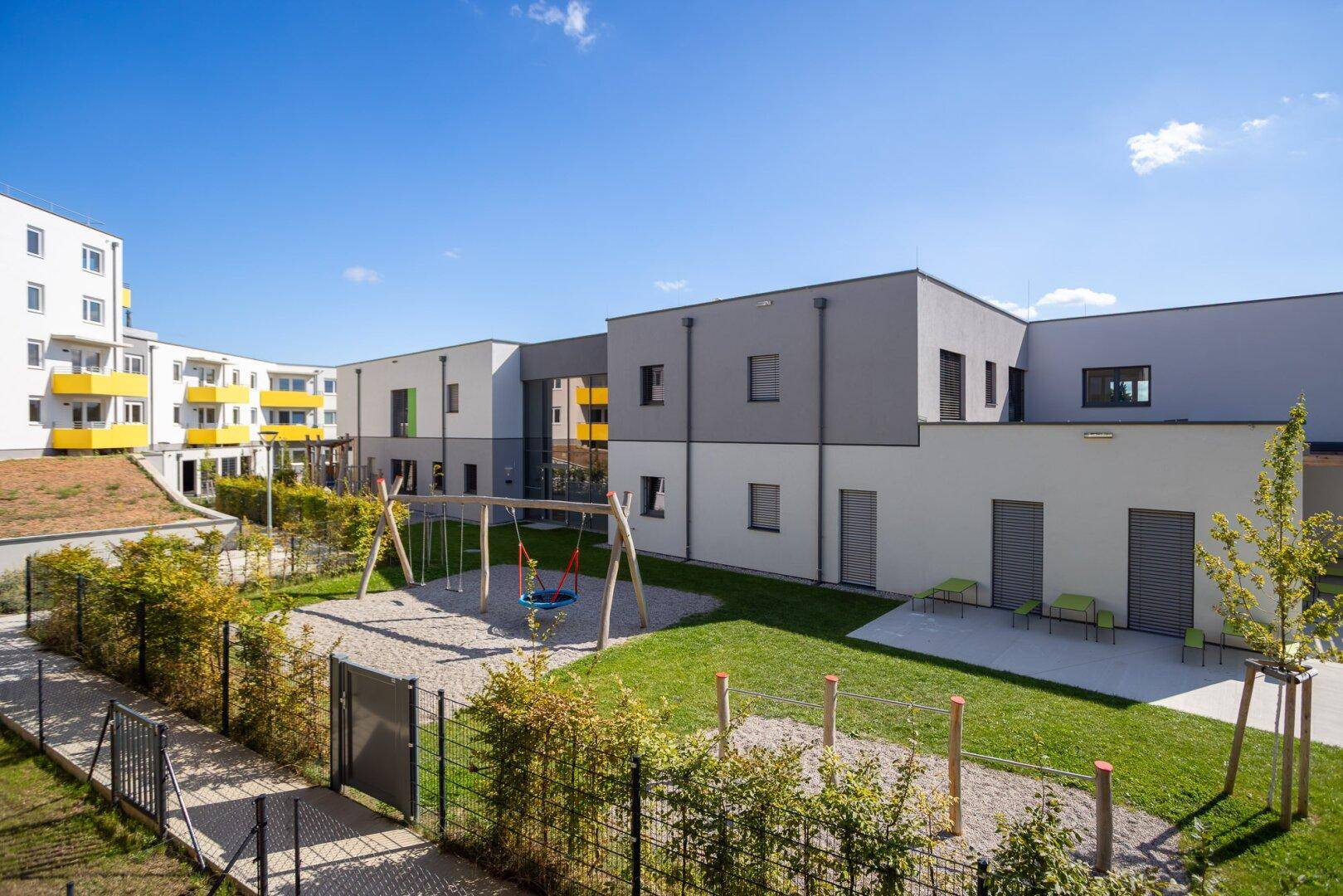 Weitläufige Grünfläche mit Spielgeräten und modernen Wohngebäuden unter blauem Himmel.