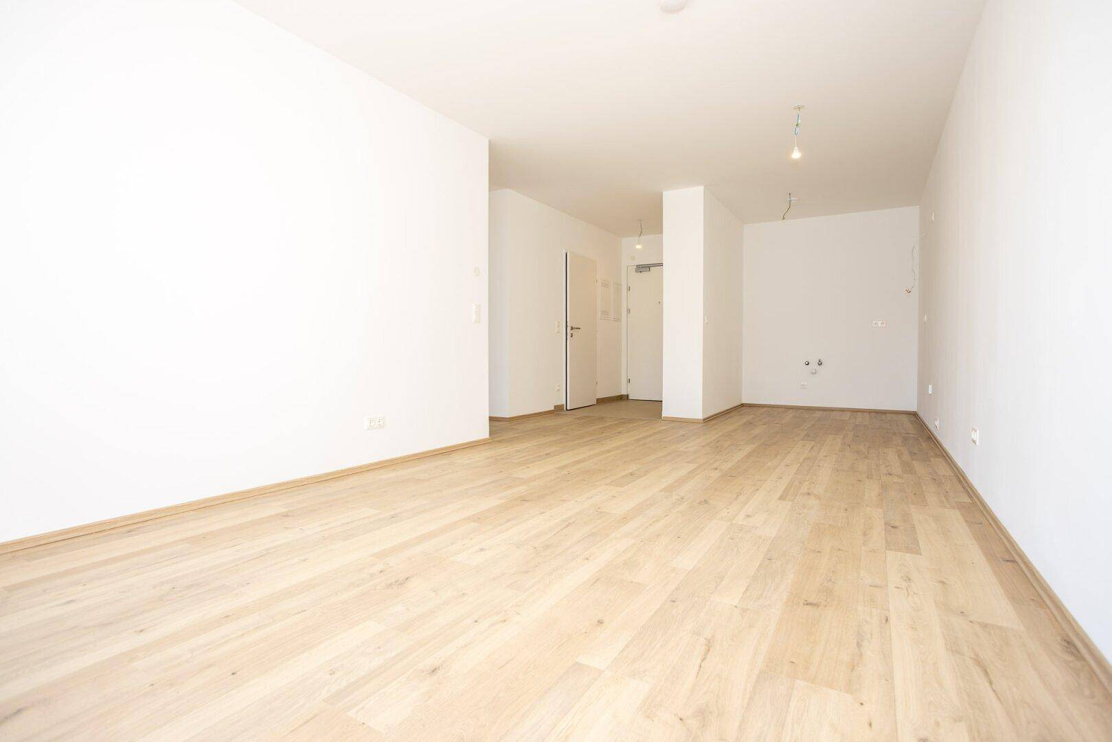 Heller, leerer Raum mit Holzboden und Zugang zum Eingangsbereich der Wohnung.