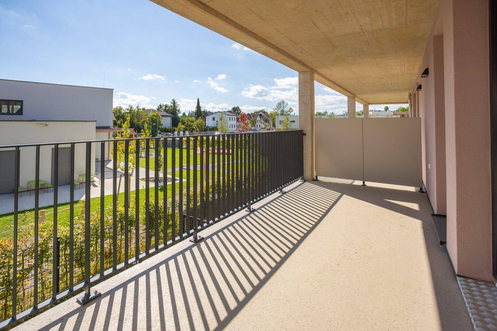Langer Balkon mit Betonboden und Blick auf eine grüne Außenanlage mit Bäumen.