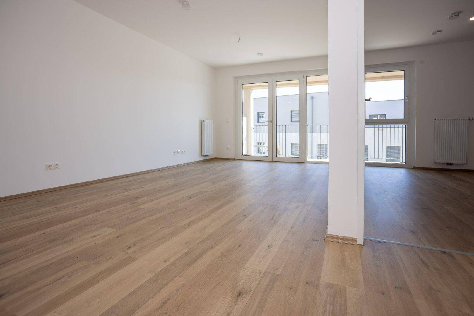 Geräumiges, leeres Zimmer mit Holzboden und großen Fenstern, die viel Licht hereinlassen.