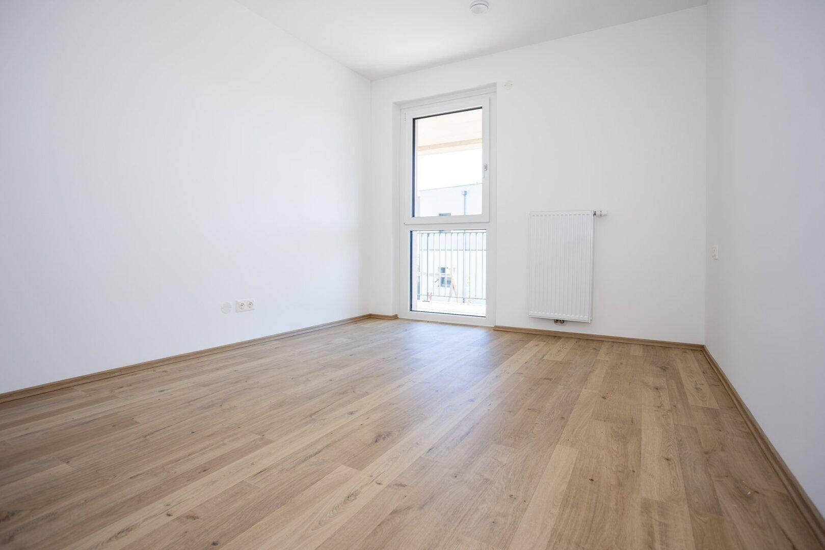 Helles Zimmer mit Holzboden und einem Fenster, das den Blick nach draußen freigibt.