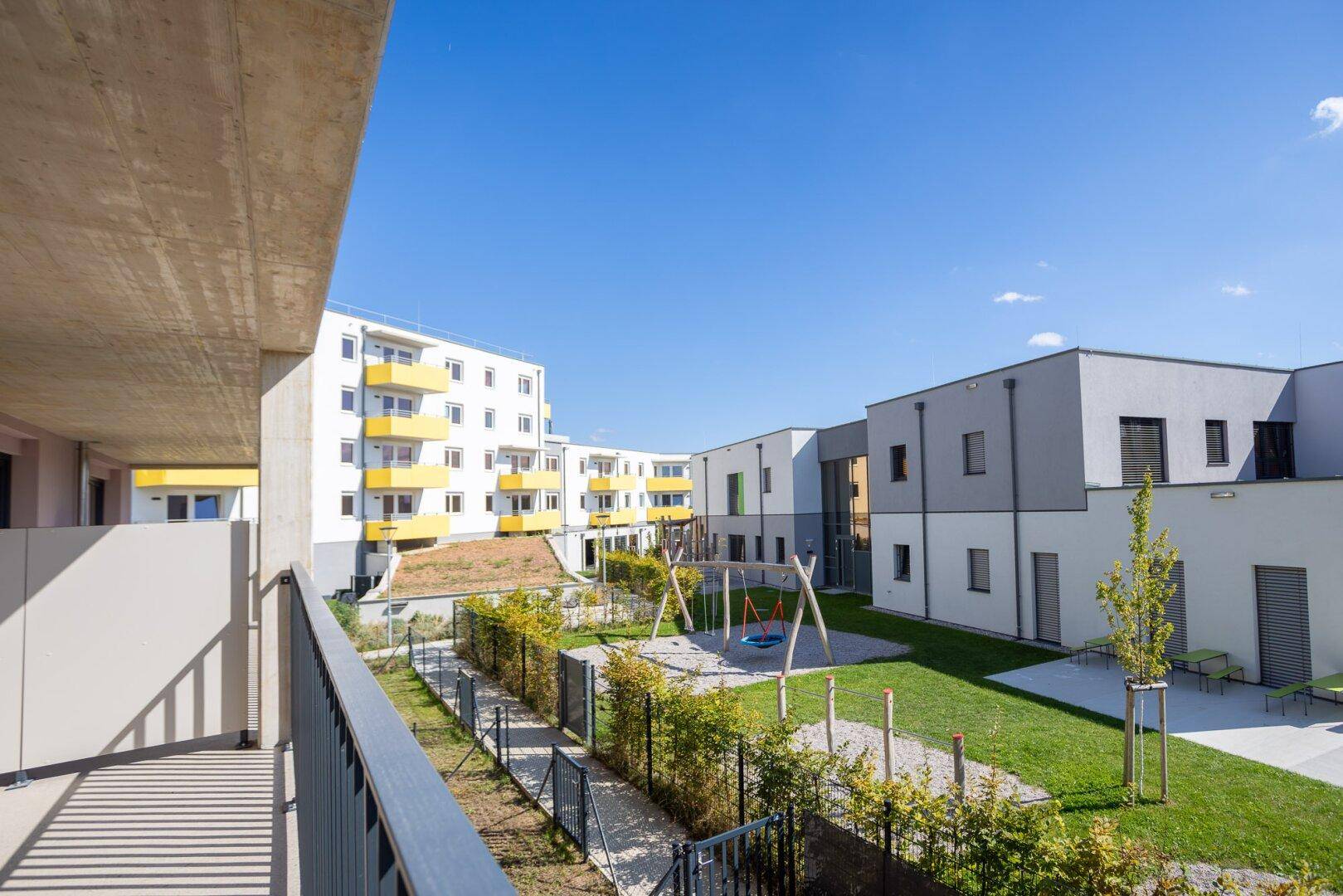 Balkon mit weitem Blick auf den grünen Innenhof mit Spielplatz und moderne Gebäude.