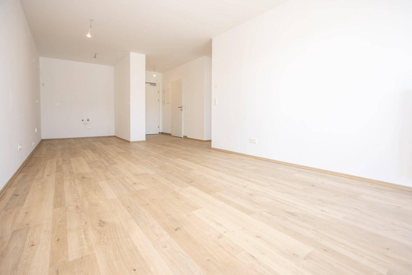 Heller Wohnbereich mit Laminatboden, weißen Wänden und Blick zum Eingangsbereich.