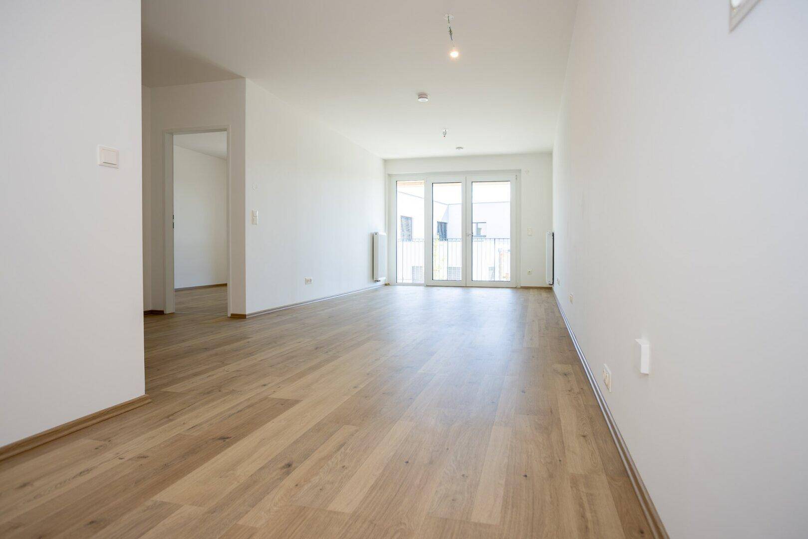 Helles, leeres Wohnzimmer mit modernem Laminatboden und Zugang zu einem Balkon.
