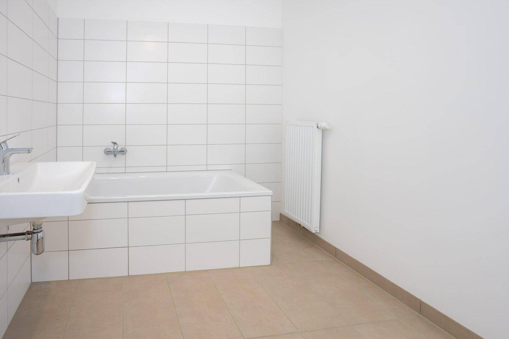 Helles Badezimmer mit weißen Fliesen, beigefarbenem Boden und Badewanne, inklusive Heizkörper.